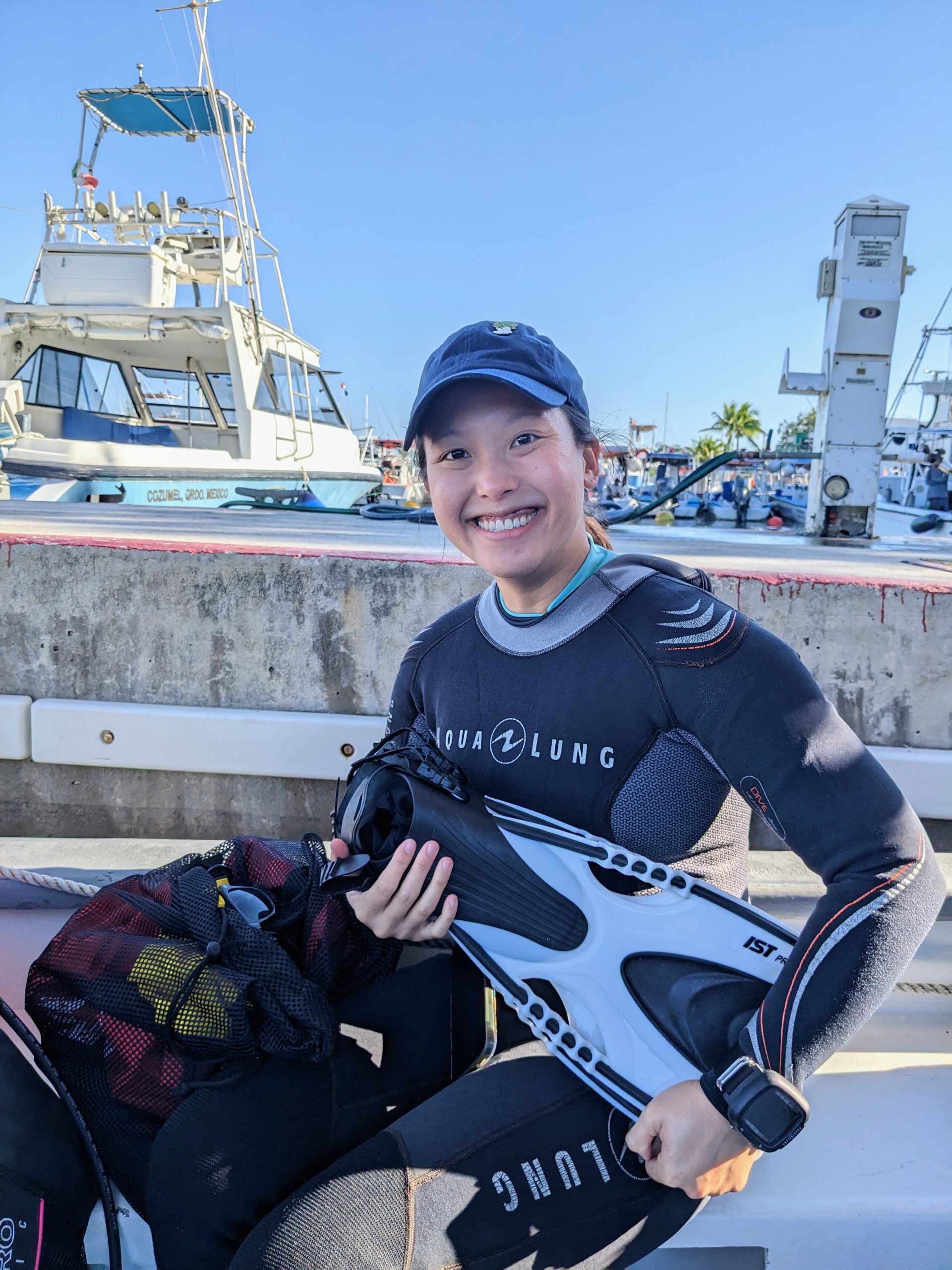 Diane at a dive port in Cozumel, wearing a full 3mm wetsuit, holding her dive fins and dive computer, and sitting next to her large bag of gear.